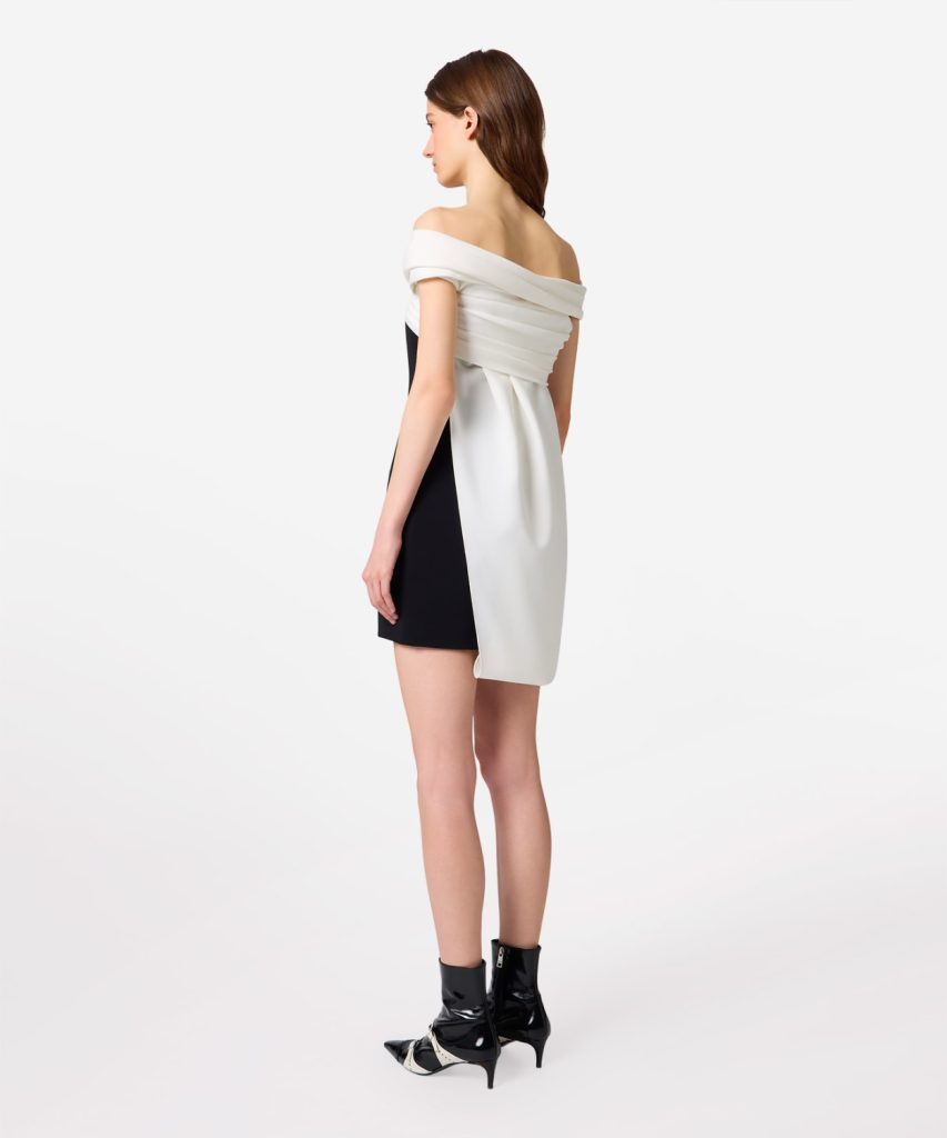 Model wearing a black and white color-block dress with an off-the-shoulder white draped top and black side panel, paired with black ankle boots.