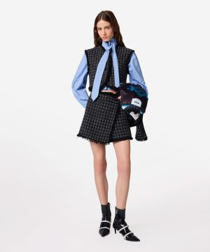 Model wearing a blue striped shirt with a black tweed vest and matching shorts, tying a blue striped scarf and holding a patterned jacket.