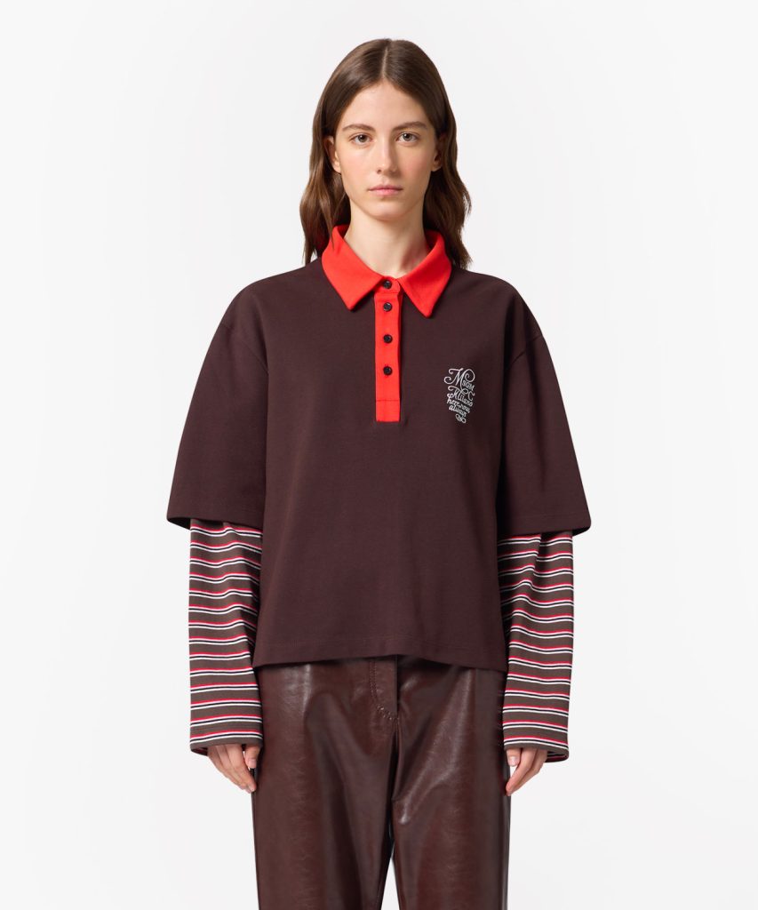Model wearing a dark brown polo with a bright red collar and placket, striped long-sleeve layer underneath, and brown leather pants.