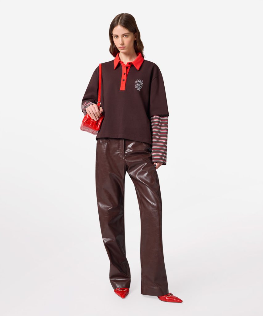 Fashion model wearing a burgundy polo with a red collar and placket, striped long sleeves, shiny brown leather pants, and red handbag and shoes.