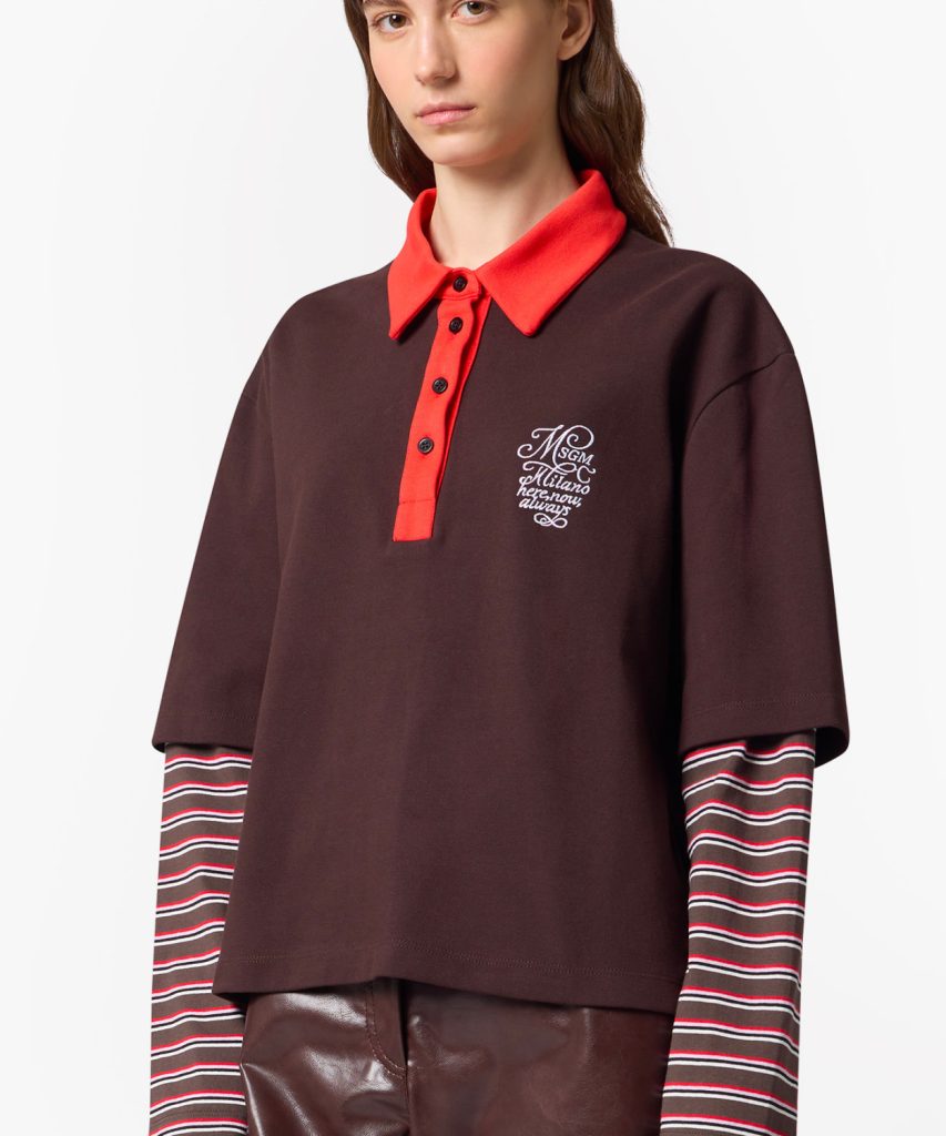 Model wearing a dark maroon polo with a bright red collar and placket, featuring a small white embroidered logo on the chest; striped long sleeves underneath and brown pants visible.