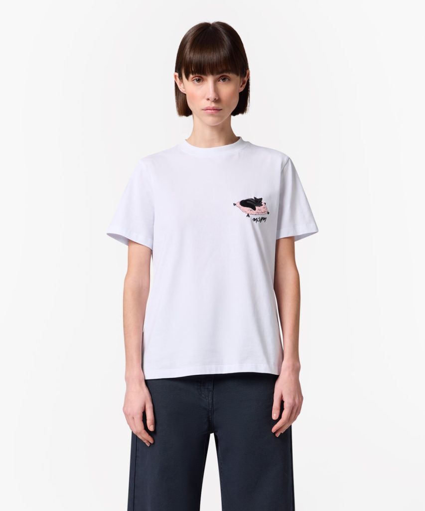 Model wearing a white T-shirt with a small embroidered patch on the left chest, paired with dark navy pants.