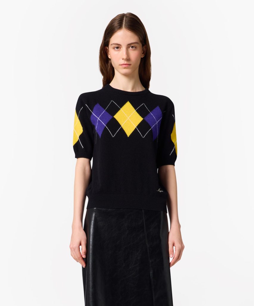 Model wearing a black knit sweater with yellow and purple argyle diamonds, paired with a black leather skirt against a white studio background.