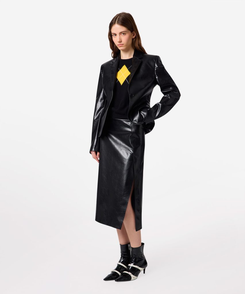 Female model wearing a black patent leather blazer and matching midi skirt with a yellow argyle sweater underneath, standing with one hand on hip.