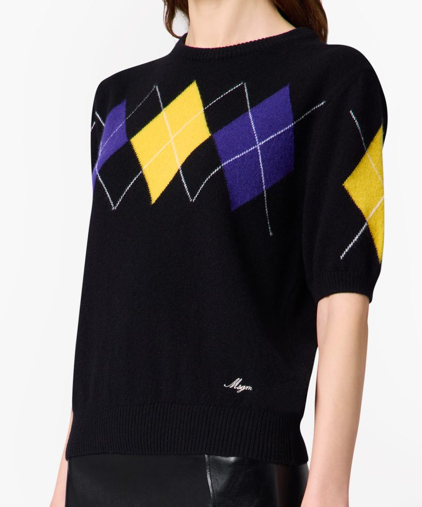 Black knit sweater featuring a multicolored argyle diamond pattern (yellow, purple) across the chest and along the sleeve with white crisscross lines, ribbed hem and short sleeves.
