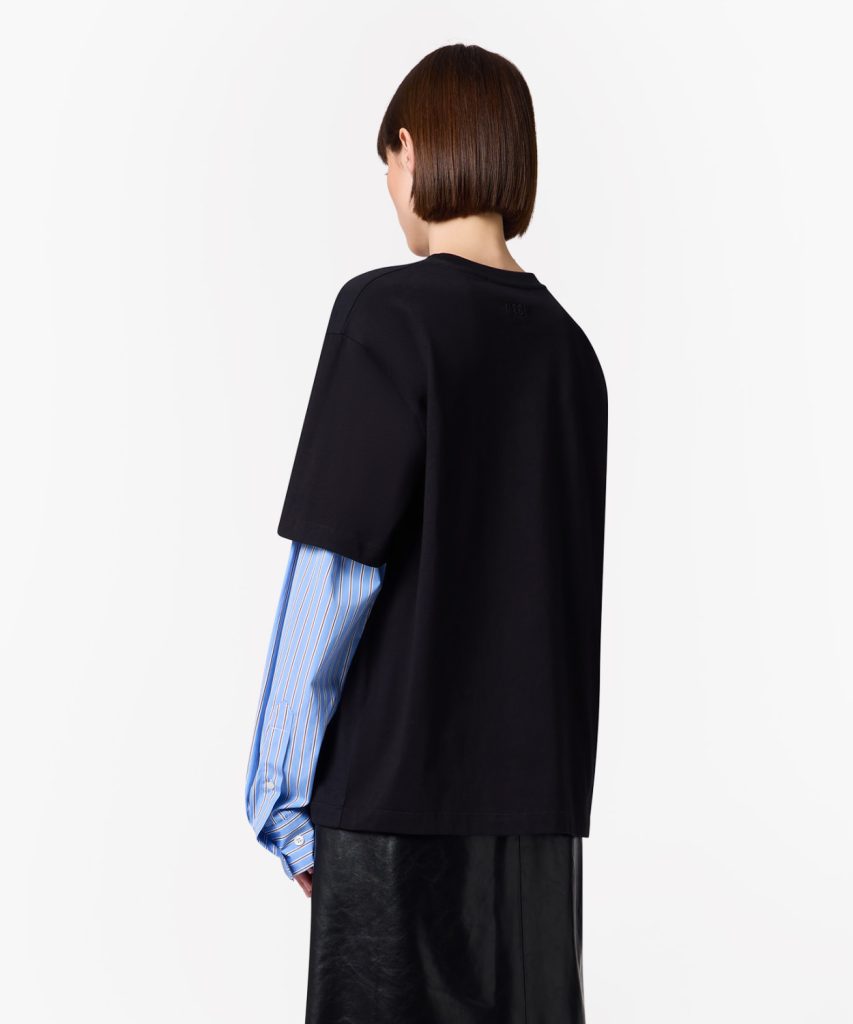Back view of a person wearing a black short-sleeve top over a blue striped shirt, standing against a white background.
