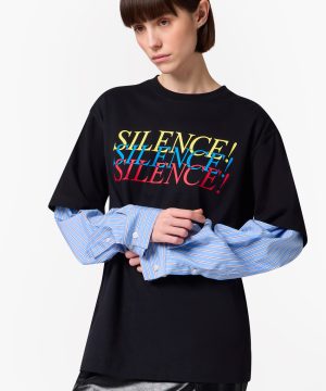 Person wearing a black T-shirt with a colorful SILENCE! graphic over a blue striped shirt, arms crossed in a fashion pose.