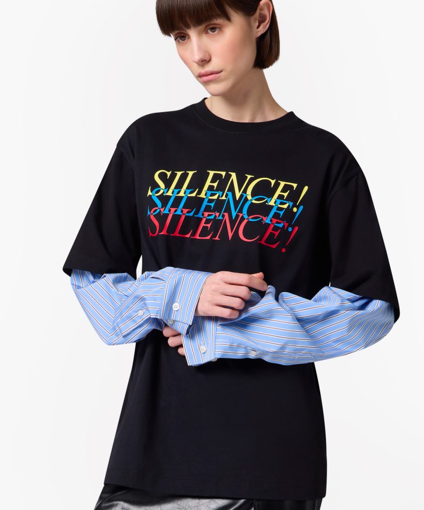 Person wearing a black T-shirt with a colorful SILENCE! graphic over a blue striped shirt, arms crossed in a fashion pose.
