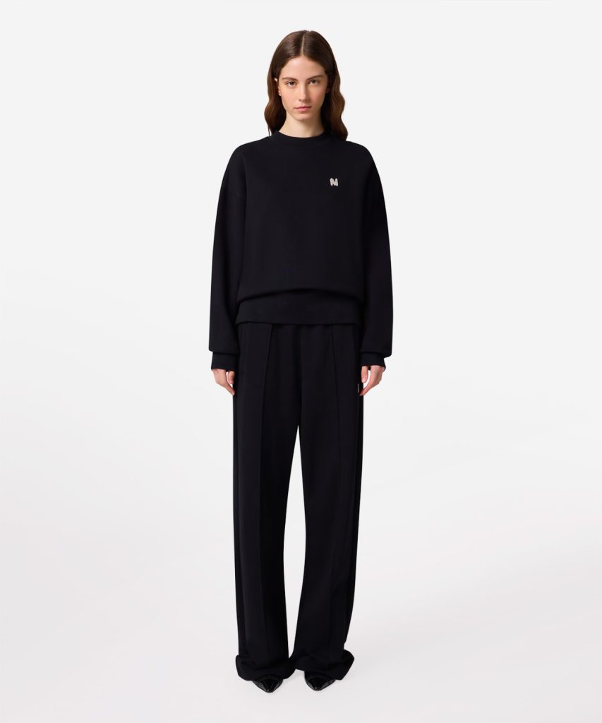 Model wearing an oversized black sweatshirt with a small logo, paired with wide-leg black trousers.