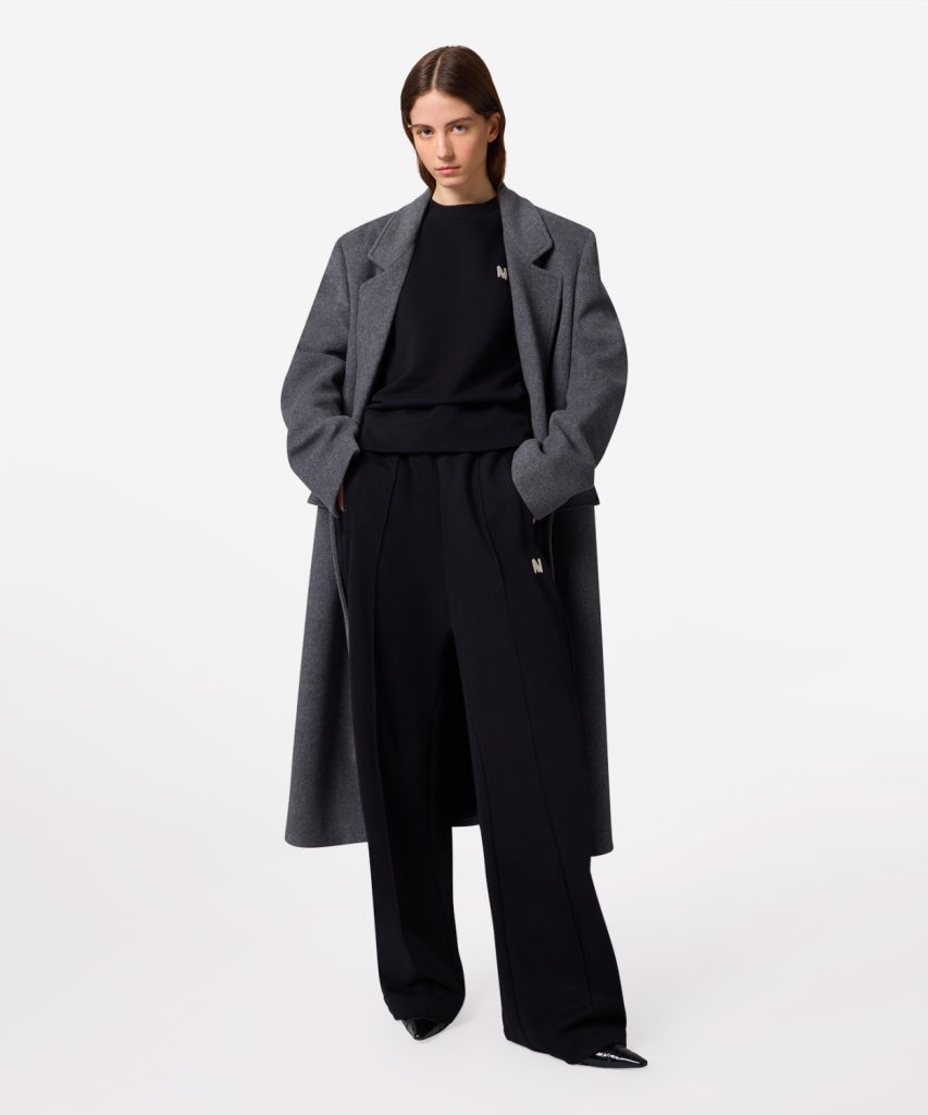Model wearing a long gray coat over a black outfit, posing with hands in pockets in a studio setting.