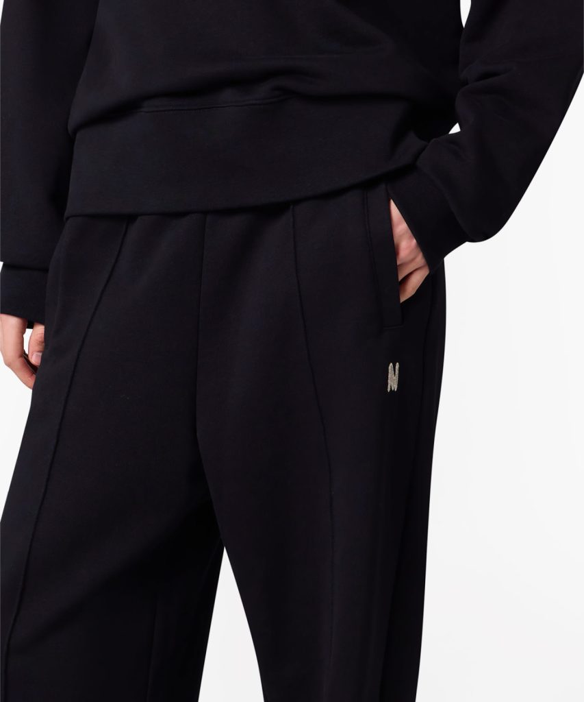 Close-up of black pants with a hand in the pocket and a small metallic 'M' logo on the hip area.
