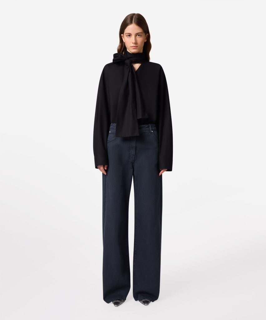 Model wearing a black blouse with a neck bow and wide-leg dark jeans against a white background.