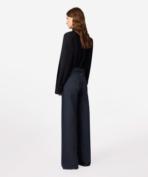 Back view of a woman wearing a black blouse and dark wide-leg jeans against a white background.