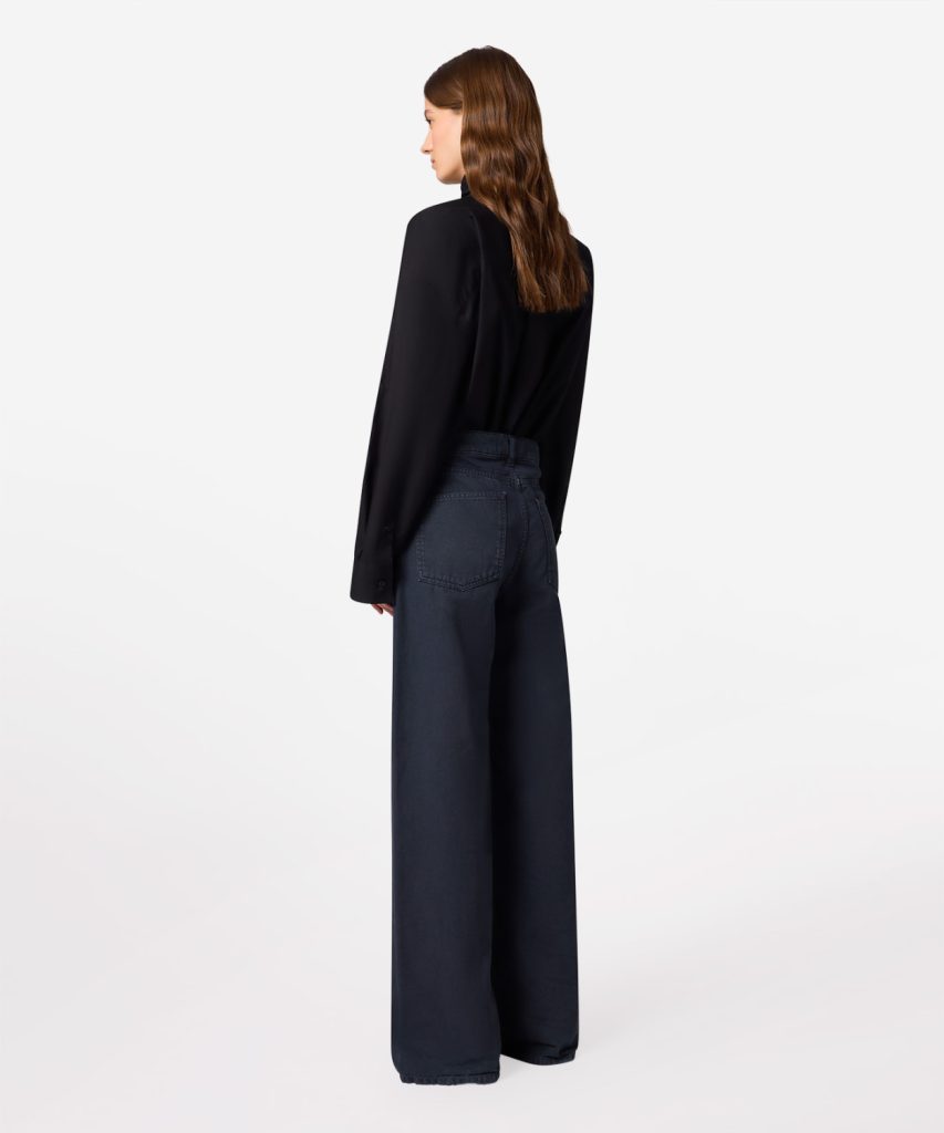 Back view of a woman wearing a black blouse and dark wide-leg jeans against a white background.