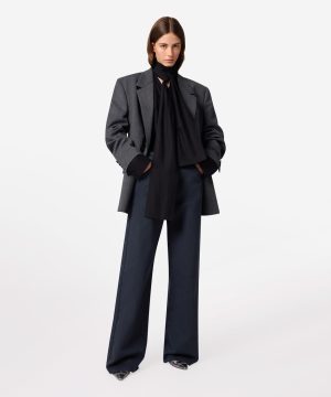 Model wearing an oversized gray blazer over a black blouse with a long scarf, paired with navy wide-leg trousers, hands in pockets.