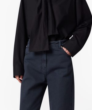 Close-up of a person wearing a black blouse and dark navy trousers with one hand in the pocket.
