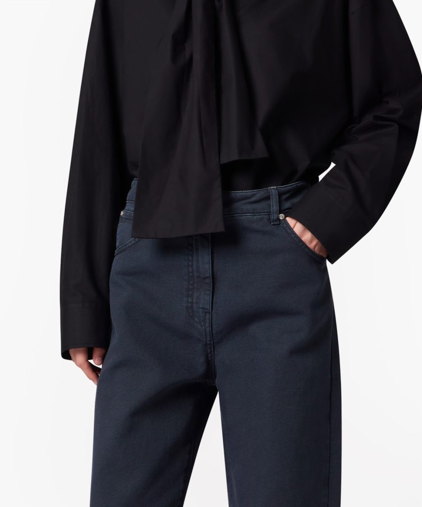 Close-up of a person wearing a black blouse and dark navy trousers with one hand in the pocket.