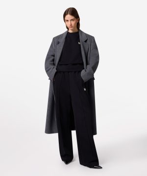 Model wearing a long gray wool coat over a black sweatshirt and wide-leg black trousers, standing with hands in pockets.