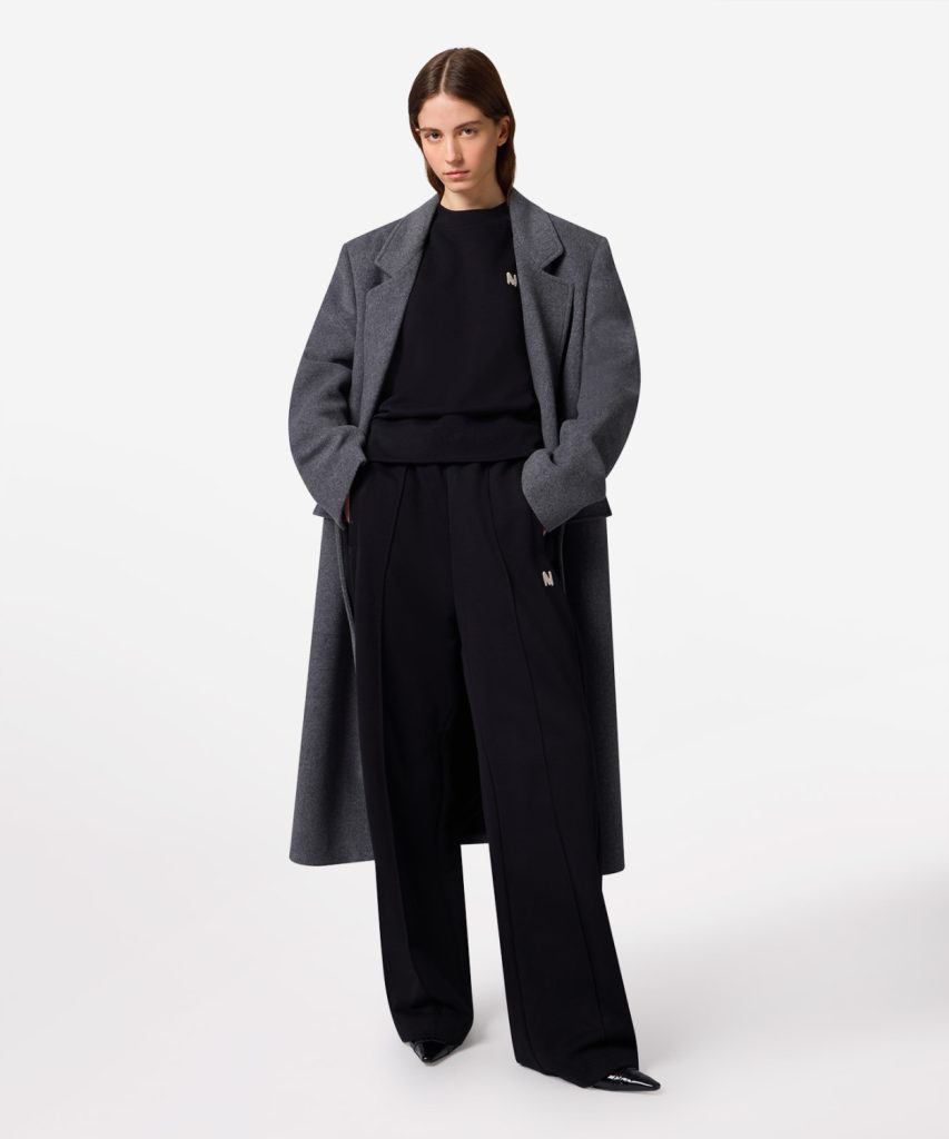 Model wearing a long gray wool coat over a black sweatshirt and wide-leg black trousers, standing with hands in pockets.