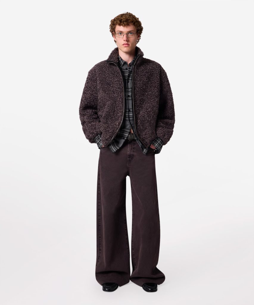 Young man modeling a dark brown fuzzy zip-up jacket over a plaid shirt with wide dark brown pants, facing the camera.