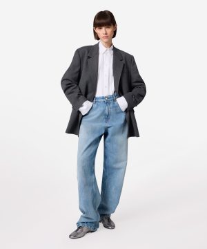 Person wearing an oversized dark blazer with a white button-down shirt and baggy blue jeans, posed with hands in pockets.