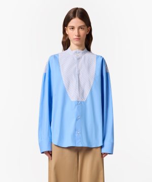 Front view of a woman wearing a light blue oversized shirt with a striped bib detail and tan wide-leg pants against a white background