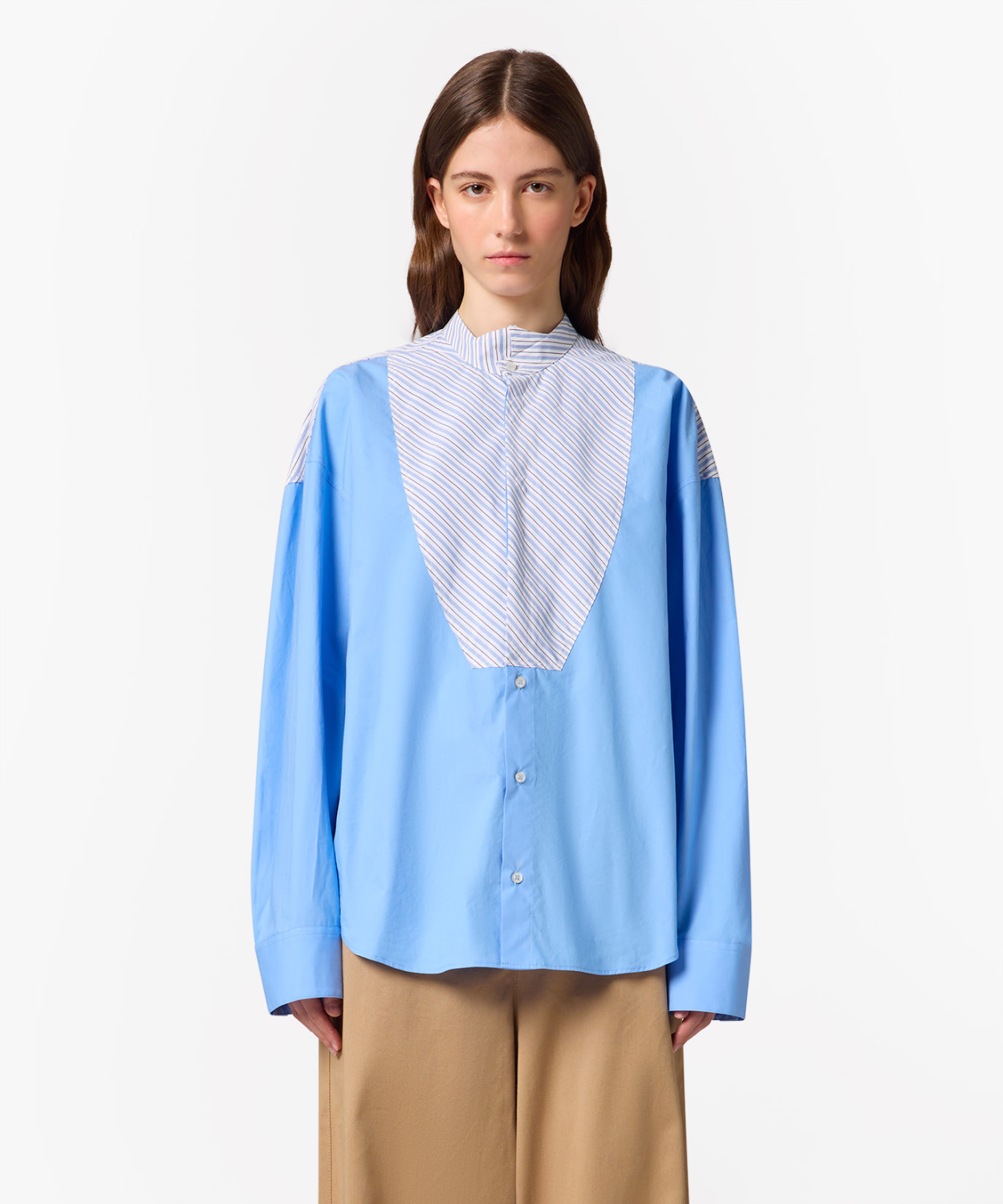 IMG_3241 - New Trend Front view of a woman wearing a light blue oversized shirt with a striped bib detail and tan wide-leg pants against a white background","Model in a blue button-down shirt with a white and blue striped front panel, standing against a white background","Person wearing a blue shirt with a V-shaped striped bib and beige trousers, facing forward","Blue long-sleeve shirt with a striped chest panel over tan pants, model looking straight ahead","Oversized blue shirt with striped front panel paired with camel-colored pants on a neutral backdrop
