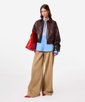 Model wearing a shiny brown leather jacket over a blue striped shirt, with wide beige pants and red accessories (bag and shoes).