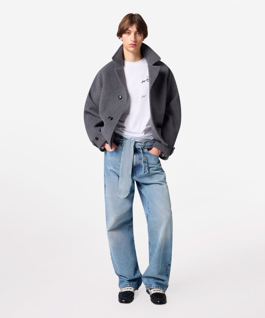 Model wearing an oversized gray coat over a white T-shirt and light-wash baggy jeans, standing with hands in pockets.