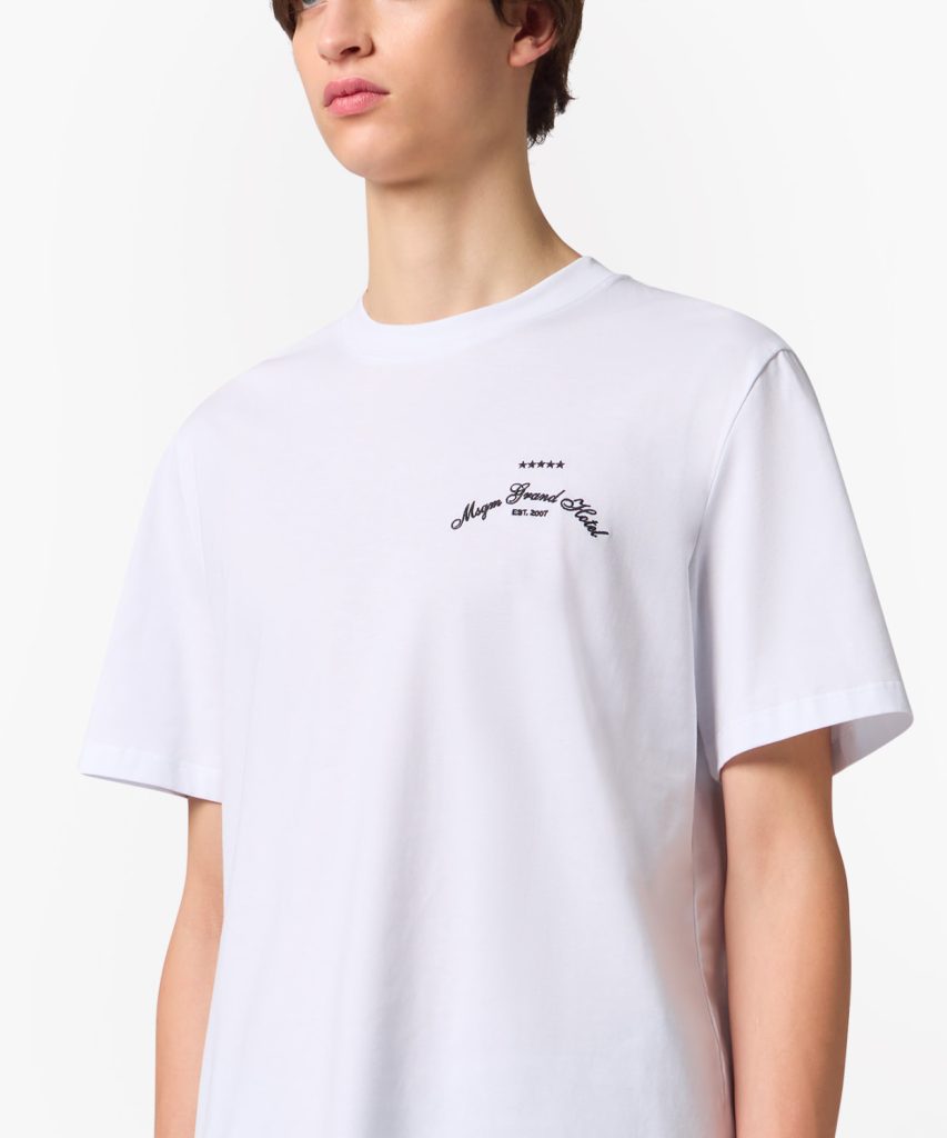 White T-shirt with a small black embroidered logo on the chest displaying cursive text and five stars above it.