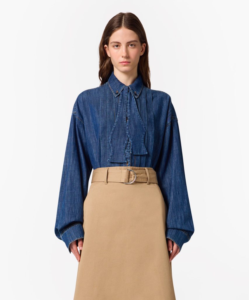 Woman wearing a blue denim blouse with a tied neck detail and a tan high-waisted skirt with a belt, neutral studio background.