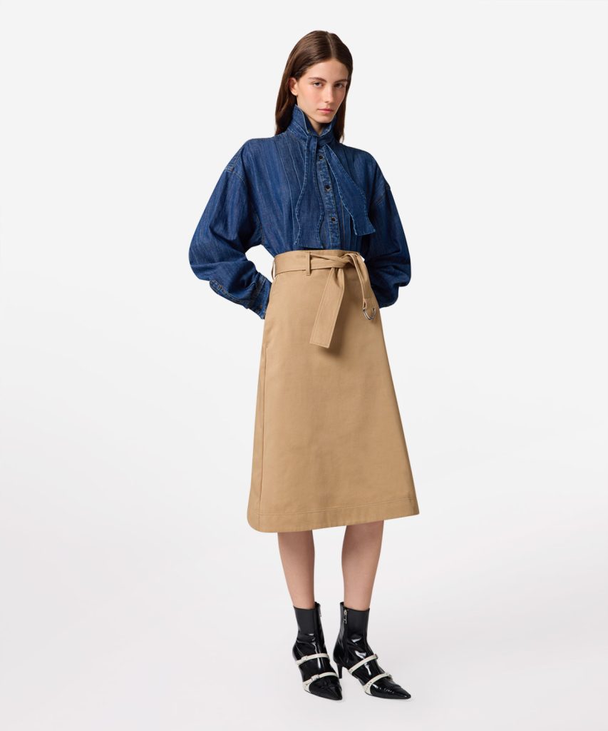 Model wearing a blue denim blouse with a high neck and puff sleeves, paired with a tan midi skirt and belt, standing against a white background.