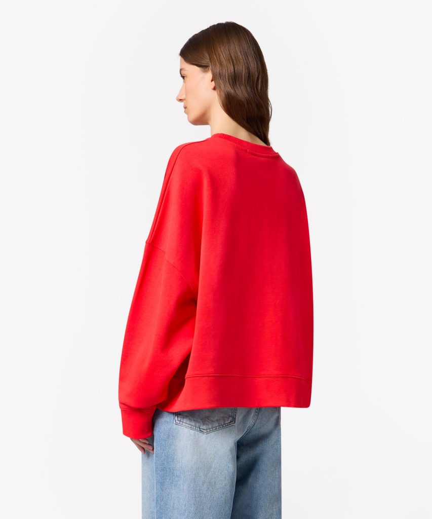 Person seen from behind wearing a bright red oversized sweatshirt with light-wash jeans, neutral background