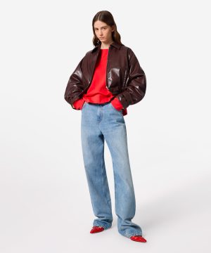 Model wearing a dark brown leather jacket over a bright red sweater and light-wash jeans, standing with hands in pockets and a relaxed pose.