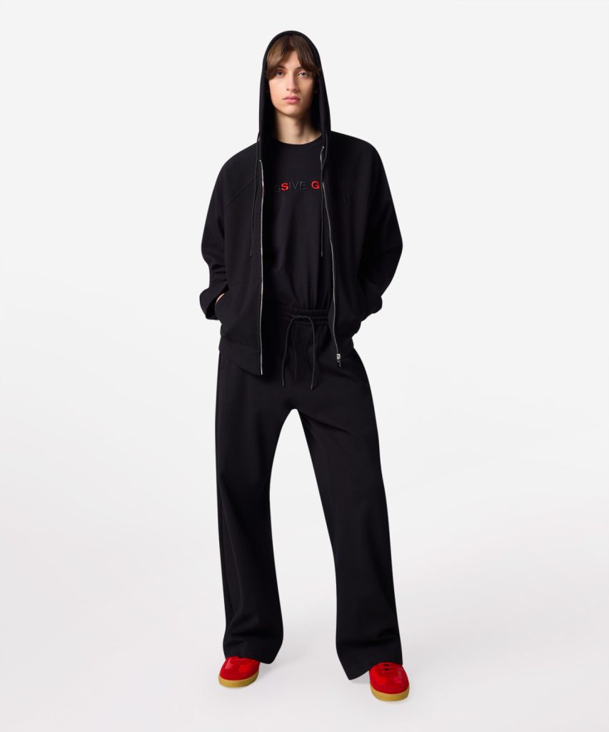 Full-body shot of a person in an all-black streetwear outfit (hoodie, sweatshirt, joggers) with red sneakers, standing with hands in pockets against a white background on a studio backdrop.