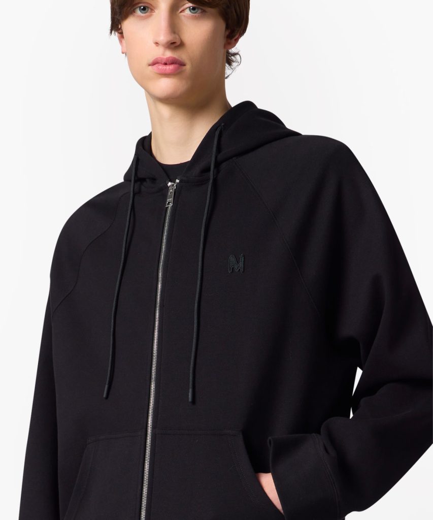 Person wearing a black zip-up hoodie with drawstrings, hands in pockets, small logo on chest.