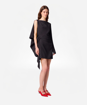 Model wearing a black asymmetrical dress with cape sleeves and a draped waist, paired with red pointed heels against a white background.