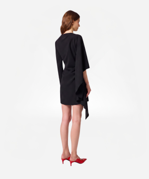 Back view of a woman wearing a knee-length black dress with an asymmetric draped hem, and red heeled shoes.