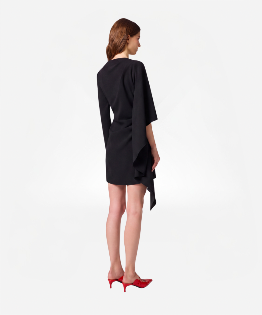 Back view of a woman wearing a knee-length black dress with an asymmetric draped hem, and red heeled shoes.