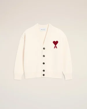 Cream cardigan with a red heart appliqué on the left chest, button-front and ribbed cuffs against a pale beige background.