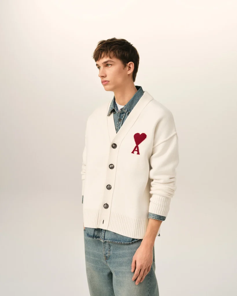 Side view of a young person wearing a cream cardigan with a red heart patch over a denim shirt and jeans, standing against a light background.
