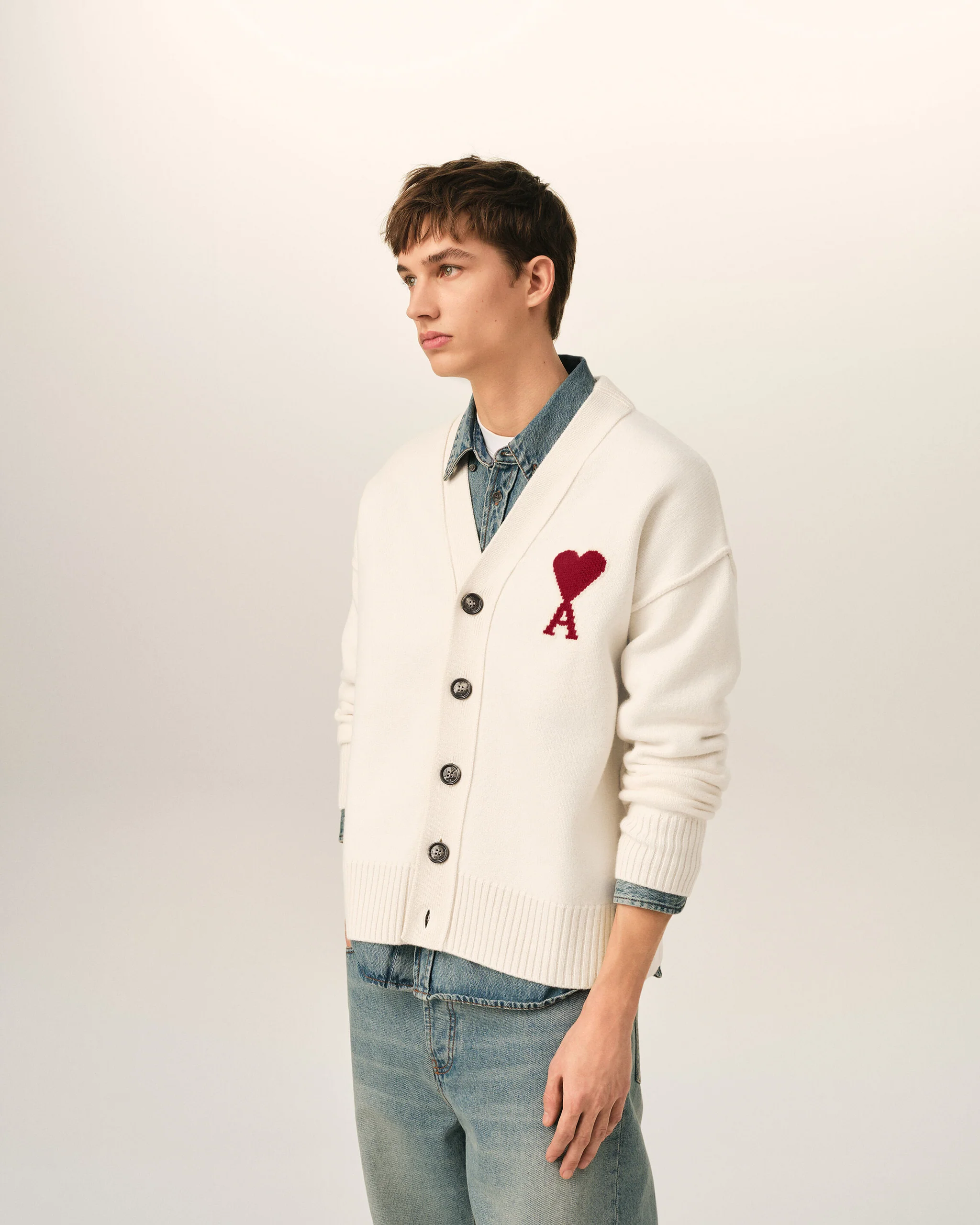 IMG_3433 - New Trend Side view of a young person wearing a cream cardigan with a red heart patch over a denim shirt and jeans, standing against a light background.