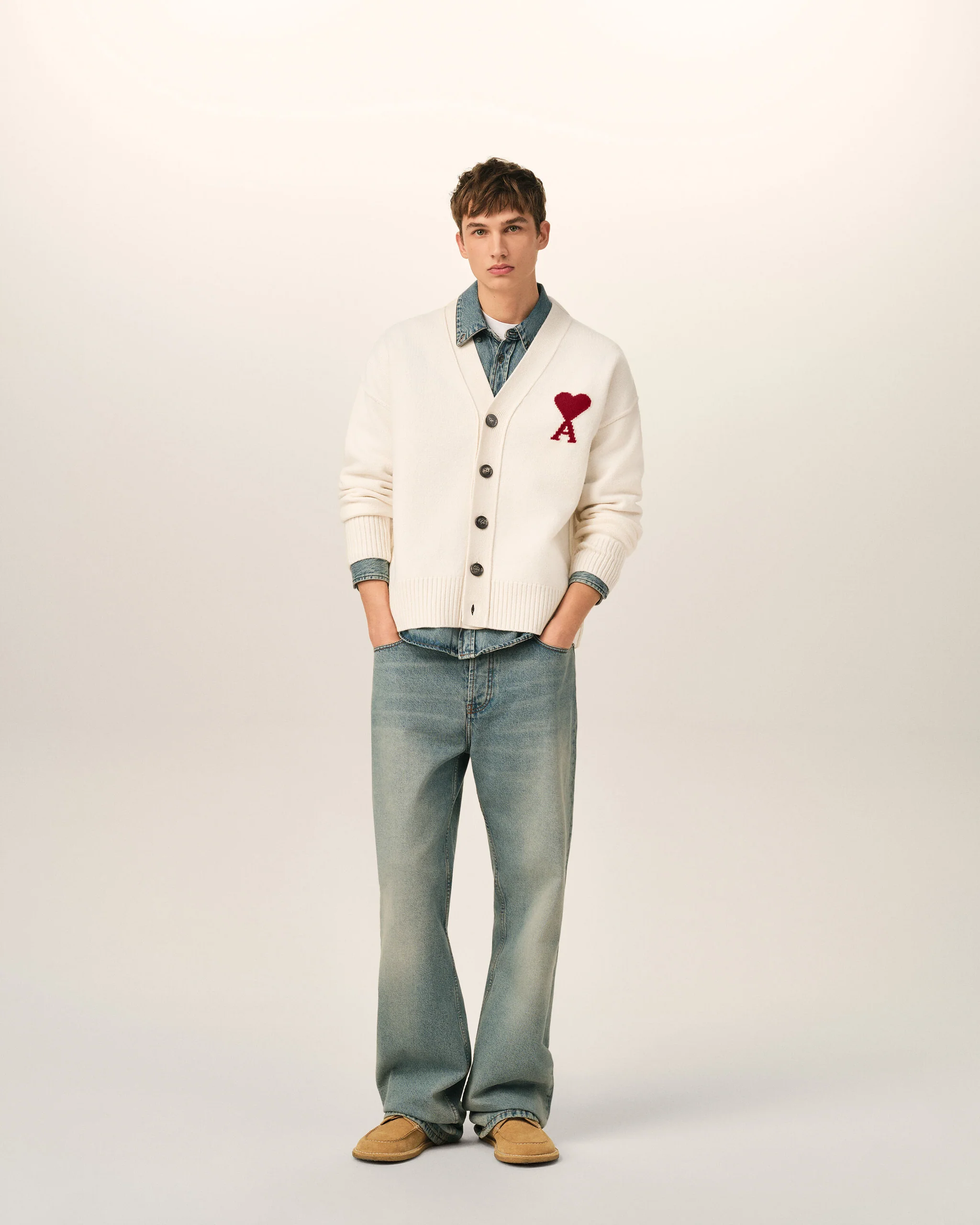 IMG_3434 - New Trend Full-body portrait of a young man in a white cardigan with a red heart patch over a denim shirt, light jeans, and tan loafers against a plain backdrop.