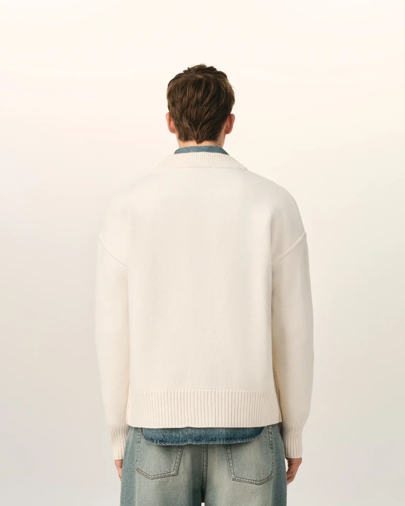 Back view of a person wearing a cream knit sweater and blue jeans against a pale background.