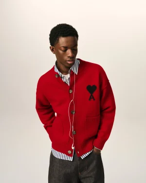 Person wearing a red cardigan with a black heart patch, striped shirt, and jeans, looking down with white earphones.