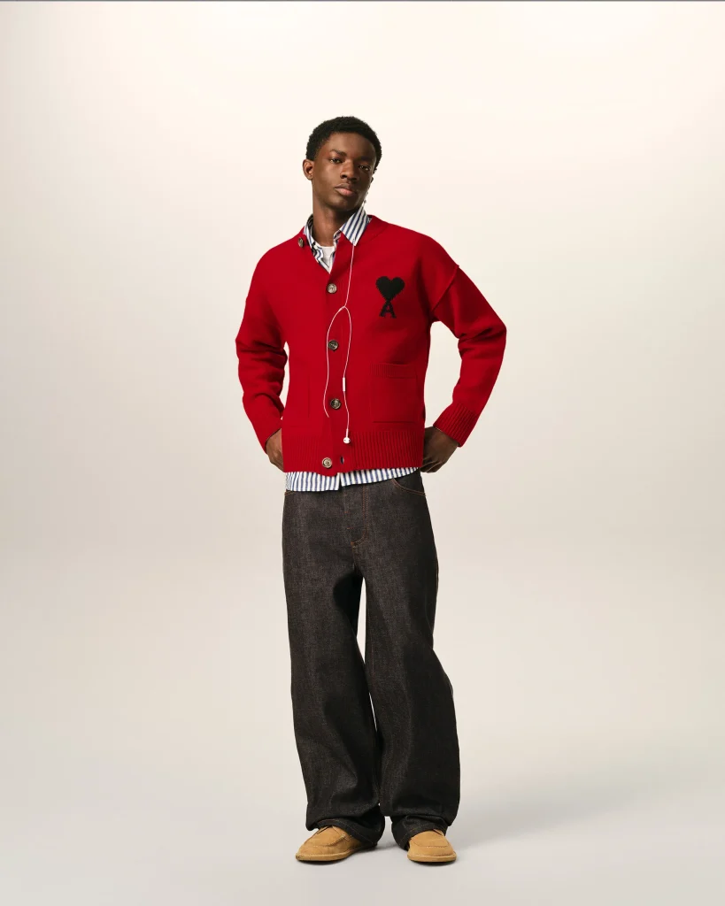 Young man posing in a red cardigan with a heart patch over a striped shirt, dark jeans, and tan shoes.