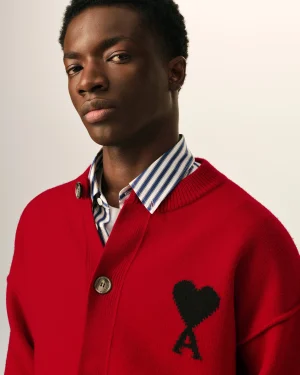 Close-up of a young man wearing a red cardigan over a blue-and-white striped shirt, with a black heart pattern on the sweater and a neutral expression.