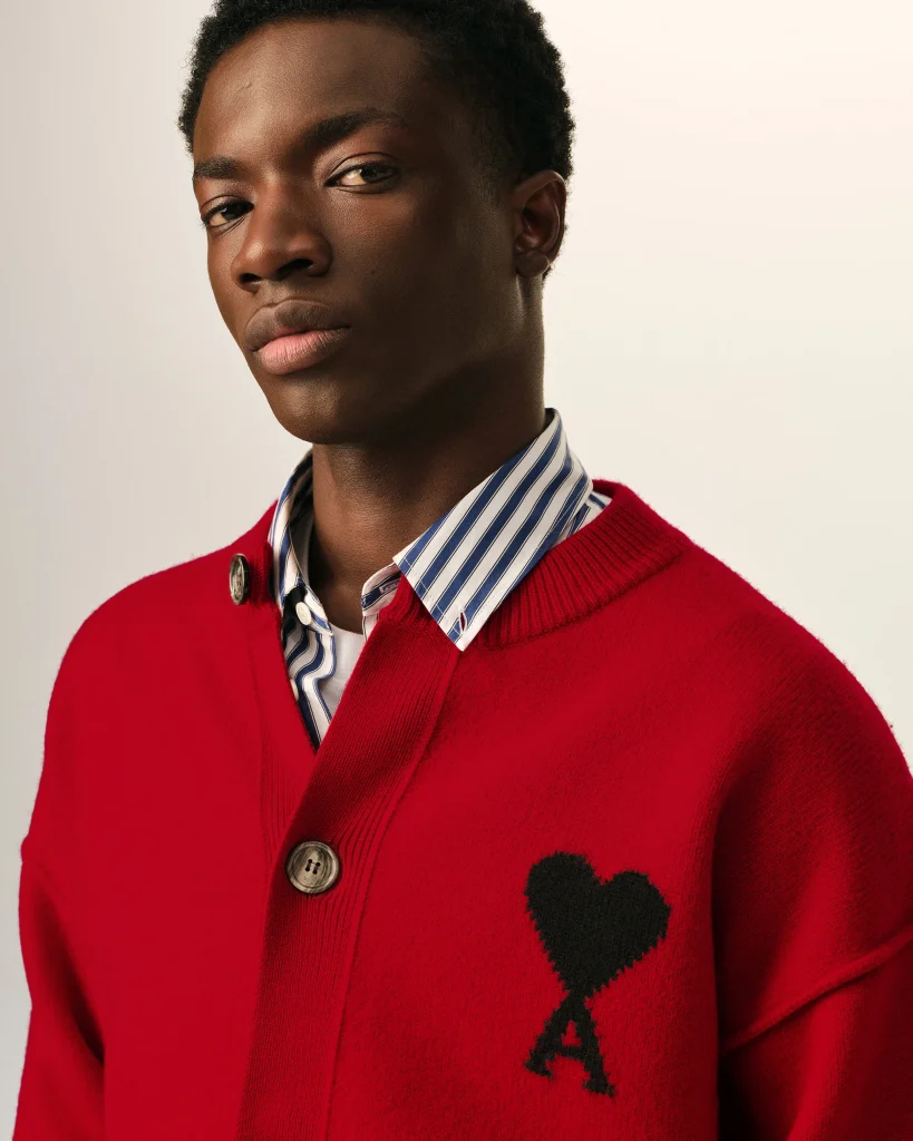 Close-up of a young man wearing a red cardigan over a blue-and-white striped shirt, with a black heart pattern on the sweater and a neutral expression.