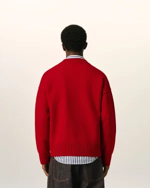 Back view of a person wearing a bright red sweater over a blue-and-white striped shirt, with dark jeans, standing against a pale background.