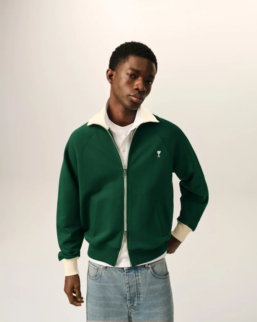 Person wearing a green zip-up jacket with a cream collar and cuffs, white shirt, and a light denim skirt against a pale background.